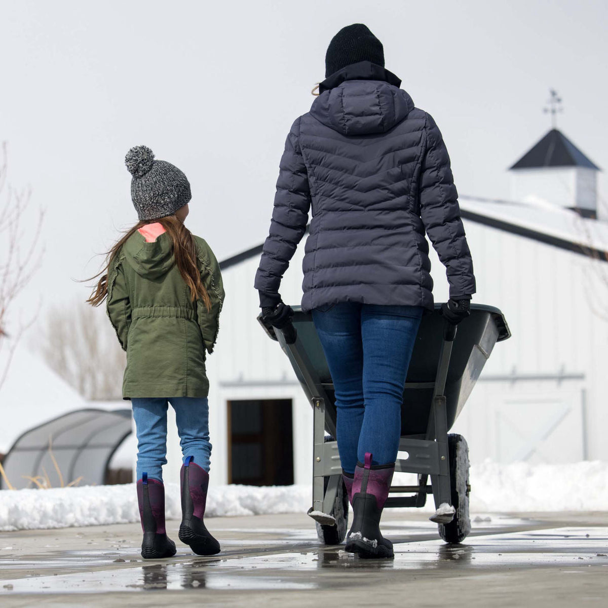 Two people pushing a wheelbarrow filled with snow in a snowy landscape.