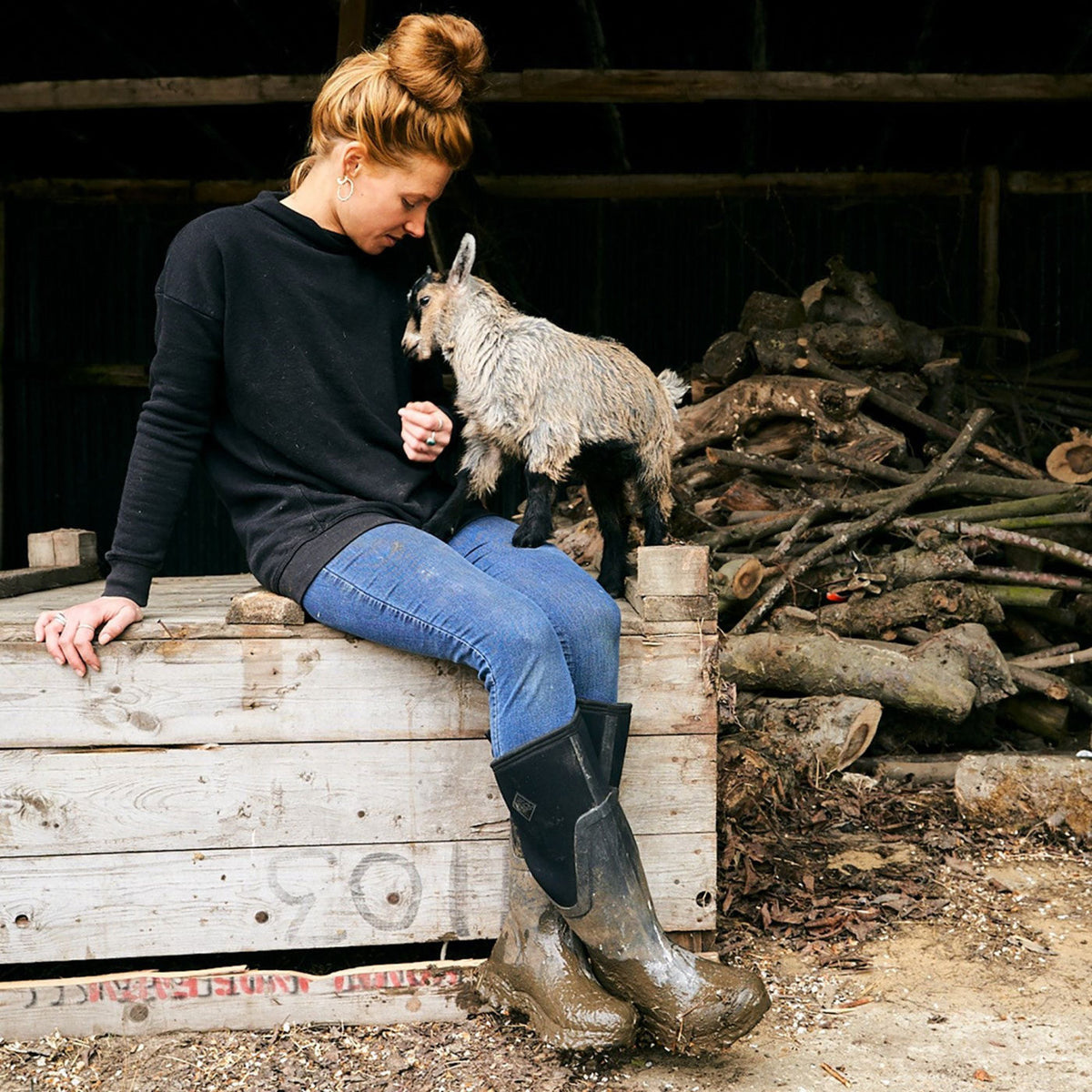 Woman sitting on a wooden crate with a small goat in a rustic setting