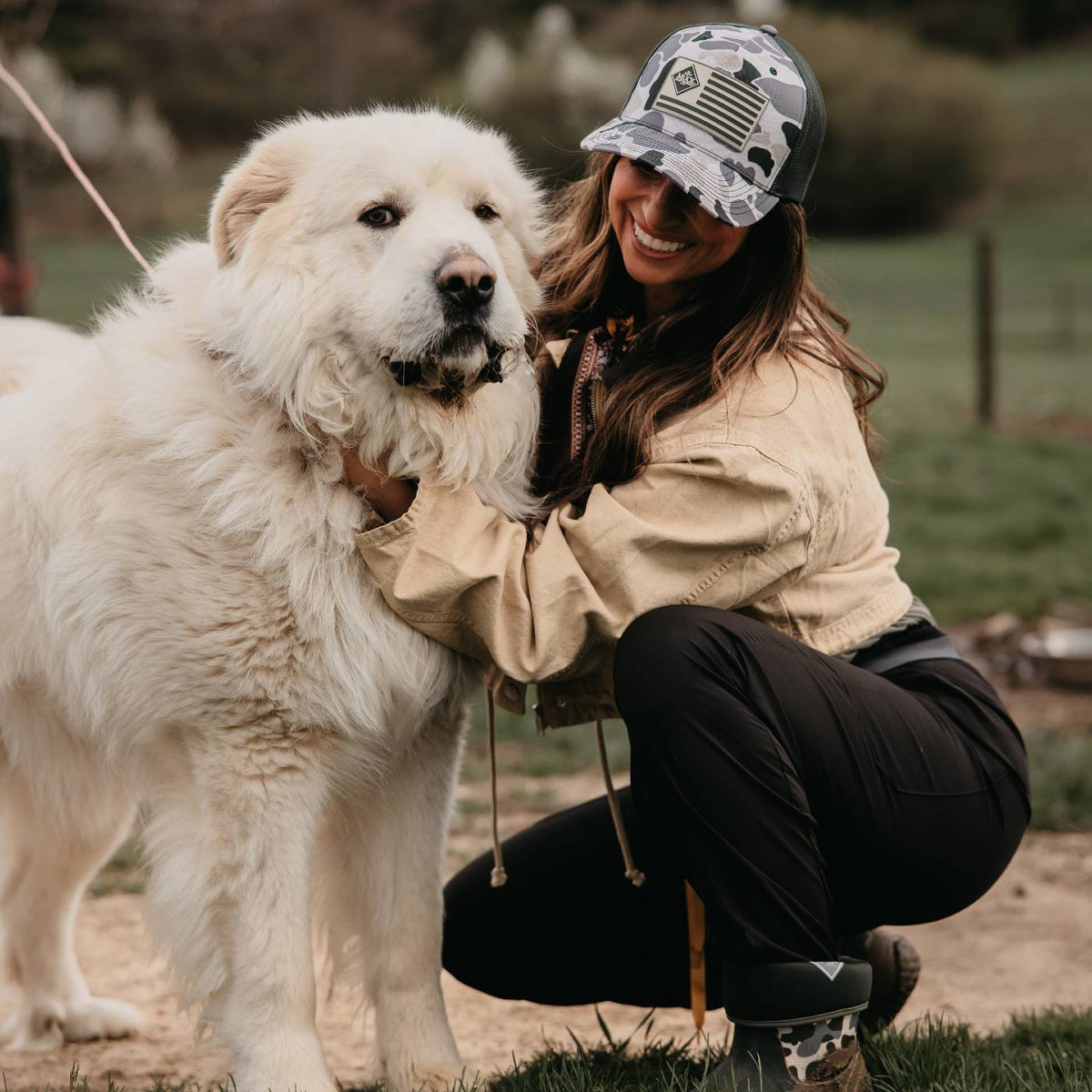 Woman with a large great pyrenees dog in a farm setting wearing a duck camo muck boots logo hat.