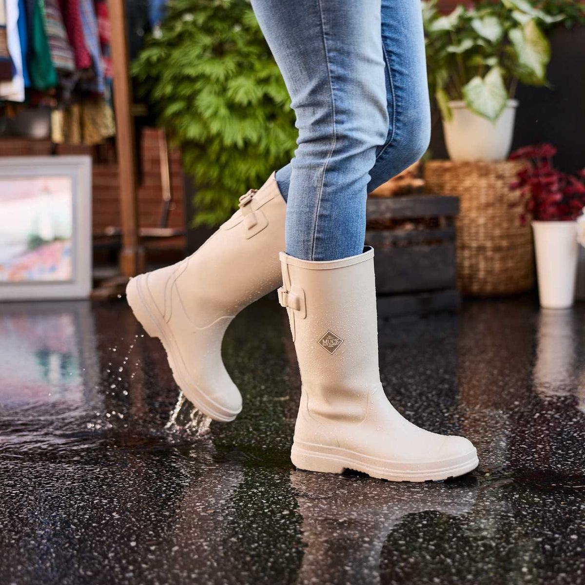 Person wearing neutral beige rubber rain boots in a a farmer's market setting with plants and clothing in the background.