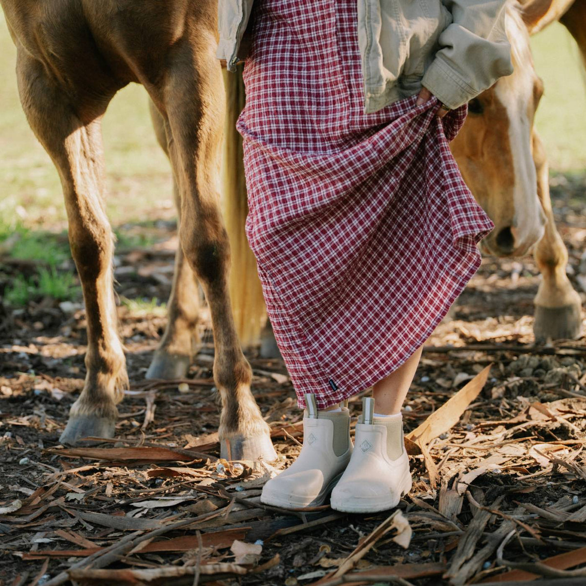 Person wearing a red plaid skirt and beige rubber chelsea boots standing next to a horse in a natural setting.