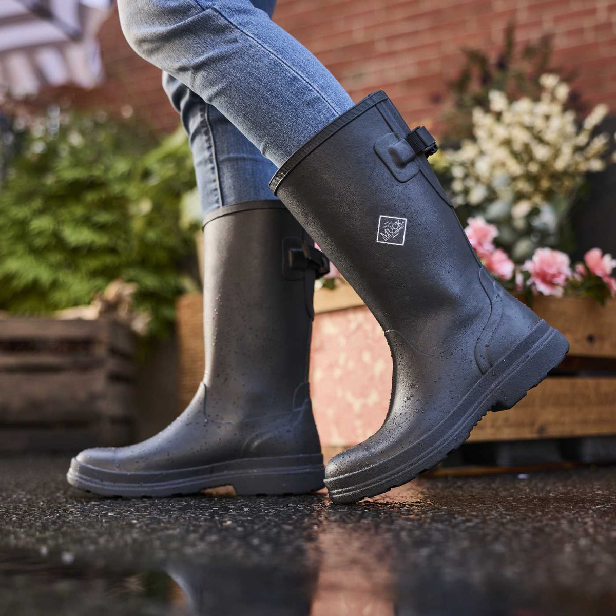 Person wearing black rubber boots with a logo, standing on a wet surface with plants in the background.