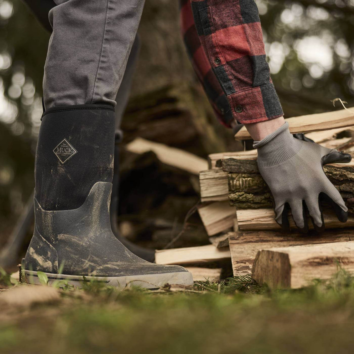 MXP12M00 Person wearing Xplorer Men's Short Waterproof Rubber Outdoor and a plaid shirt, stacking firewood outdoors.