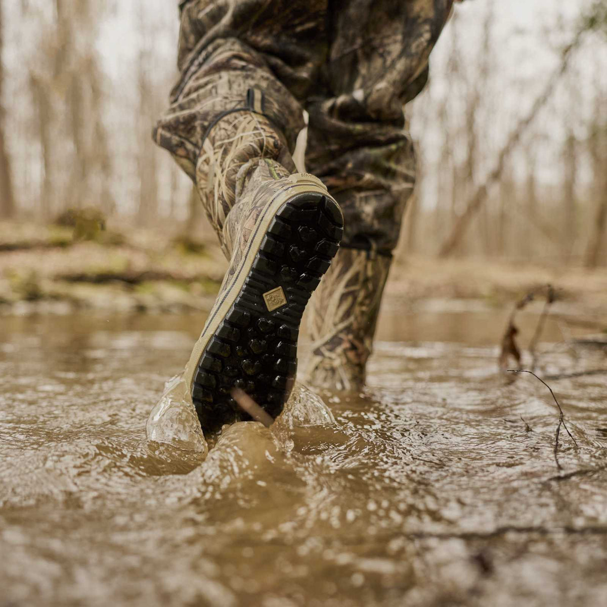 A close-up action shot of a person wading through shallow muddy water, showing the sole of a camouflage hunting boot with a black treaded outsole. The second boot is visible in the background.