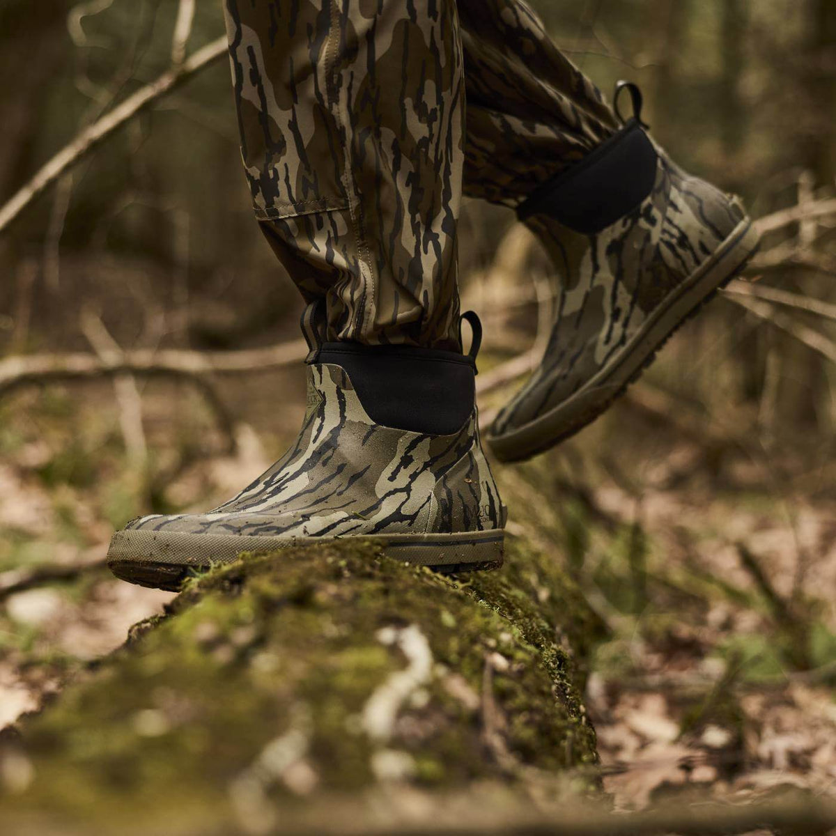 Person wearing camouflage pants and Mossy Oak Original Bottomland short waterproof rubber camouflage boots walking on a mossy rock in a forest.