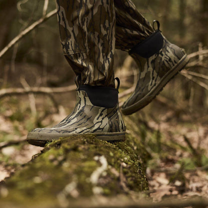 Person wearing camouflage pants and Mossy Oak Original Bottomland short waterproof rubber camouflage boots walking on a mossy rock in a forest.